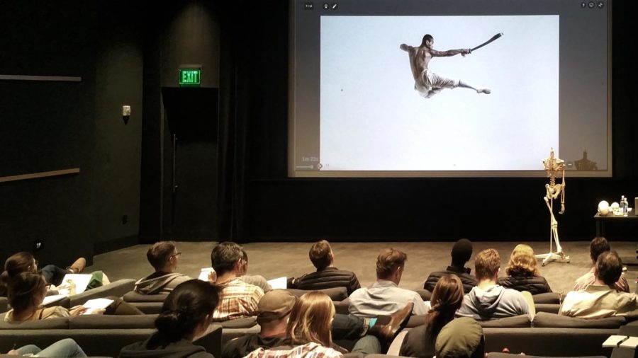 A darkened lecture theatre where a class draws from a Bodies in Motion sequence projected on the screen — a flying side-kick — beside a hanging study skeleton